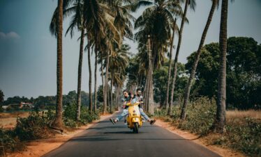 A joyful couple rides a yellow scooter through a scenic palm-lined road, capturing the essence of tropical travel and adventure.