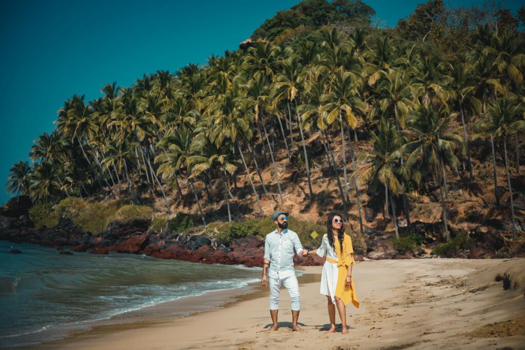 Couple enjoying a sunny beach walk in Goa with lush palm trees and clear blue waters.