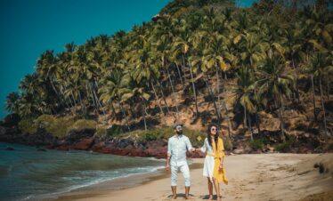 Couple enjoying a sunny beach walk in Goa with lush palm trees and clear blue waters.