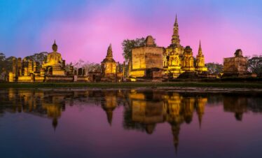 Beautiful sunset view of Wat Mahathat temple in Sukhothai, Thailand, reflecting in the surrounding water.