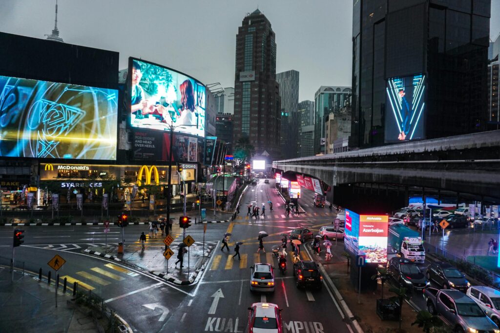 Crowded intersection in Kuala Lumpur at night, with vibrant billboards and traffic.