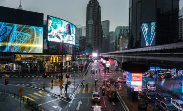 Crowded intersection in Kuala Lumpur at night, with vibrant billboards and traffic.