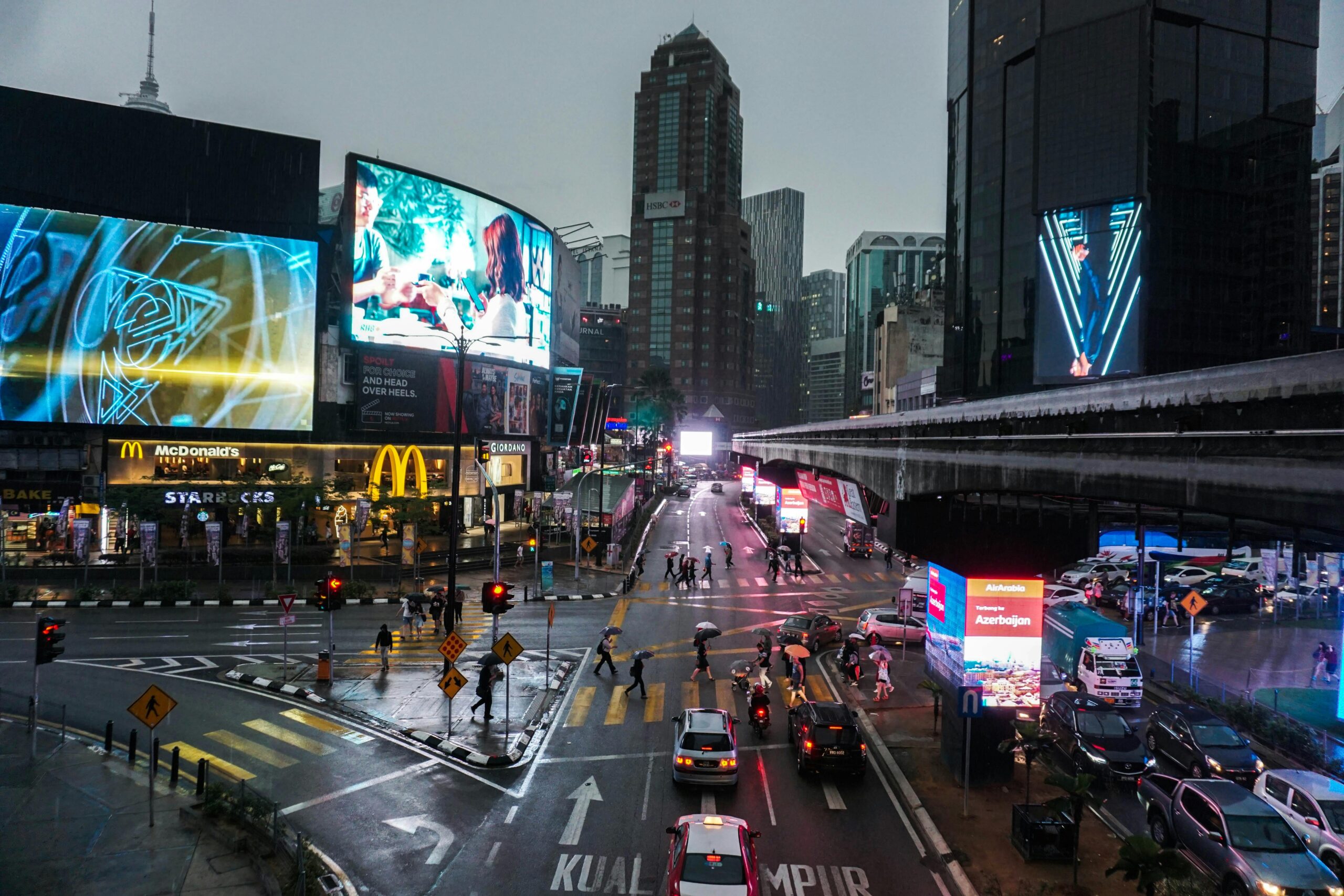Crowded intersection in Kuala Lumpur at night, with vibrant billboards and traffic.