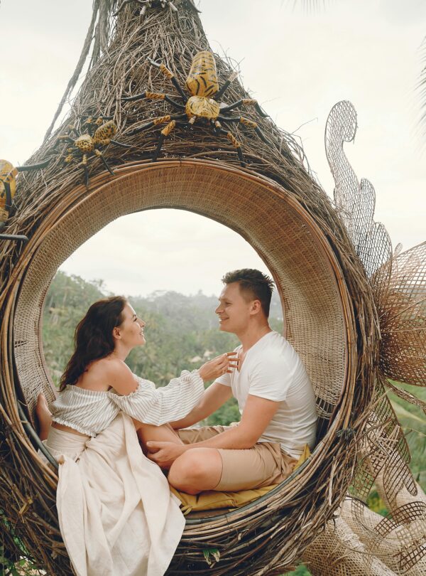 Side view full body barefoot content young couple in white casual clothes sitting in wicker hanging swing and looking at each other with love while spending honeymoon in tropical resort