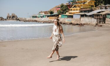 A woman happily walking on a sandy beach beside colorful coastal buildings.