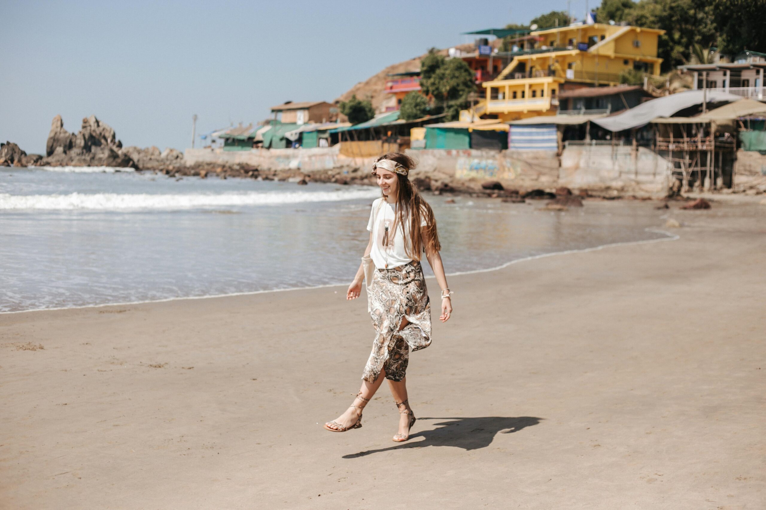 A woman happily walking on a sandy beach beside colorful coastal buildings.